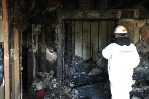 A Person In A White Protective Suit Inspects The Water-Damaged Interior Of A Building After A Fire, Including Exposed Beams And Debris.