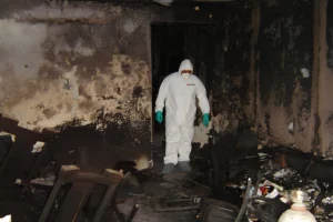 A Person In A White Protective Suit Investigates A Room Heavily Damaged By Water, With Swollen Furniture And Blackened Walls.
