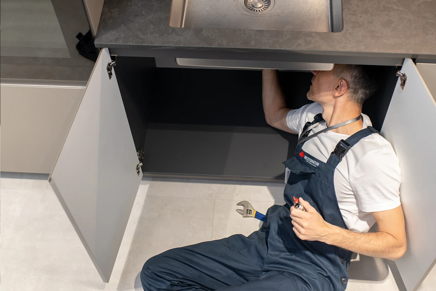 An emergency plumber lying on his back under a kitchen sink cabinet, working on plumbing. He wears a blue overall and holds tools, with cabinet doors open and sink visible above.