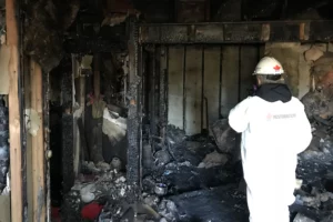 A person in a white protective suit inspects the water-damaged interior of a building after a fire, including exposed beams and debris.