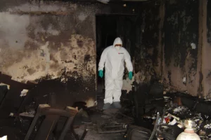 A person in a white protective suit investigates a room heavily damaged by water, with swollen furniture and blackened walls.