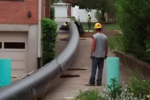 A worker in a gray t-shirt and yellow hard hat observes a large black pipe being laid along a residential driveway bordered by greenery for mold removal.