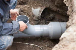 A worker in overalls connects sections of a large gray pvc pipe in a muddy excavation site, focusing on aligning the joints accurately for flood restoration.