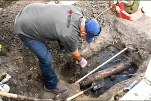 A worker in a gray shirt and blue helmet conducts a mold inspection around a large pipe in a dirt excavation site, using a shovel.